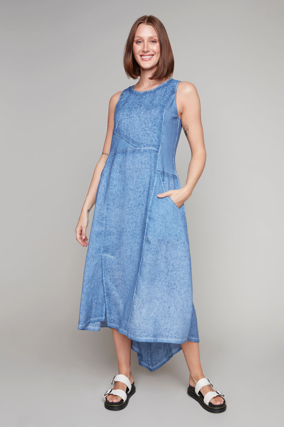 Woman wearing a sleeveless denim blue midi dress with an asymmetrical hemline and white buckle sandals.