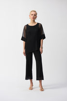 Woman wearing a black mesh cocoon top with asymmetrical hem and matching black pants, standing in full view.