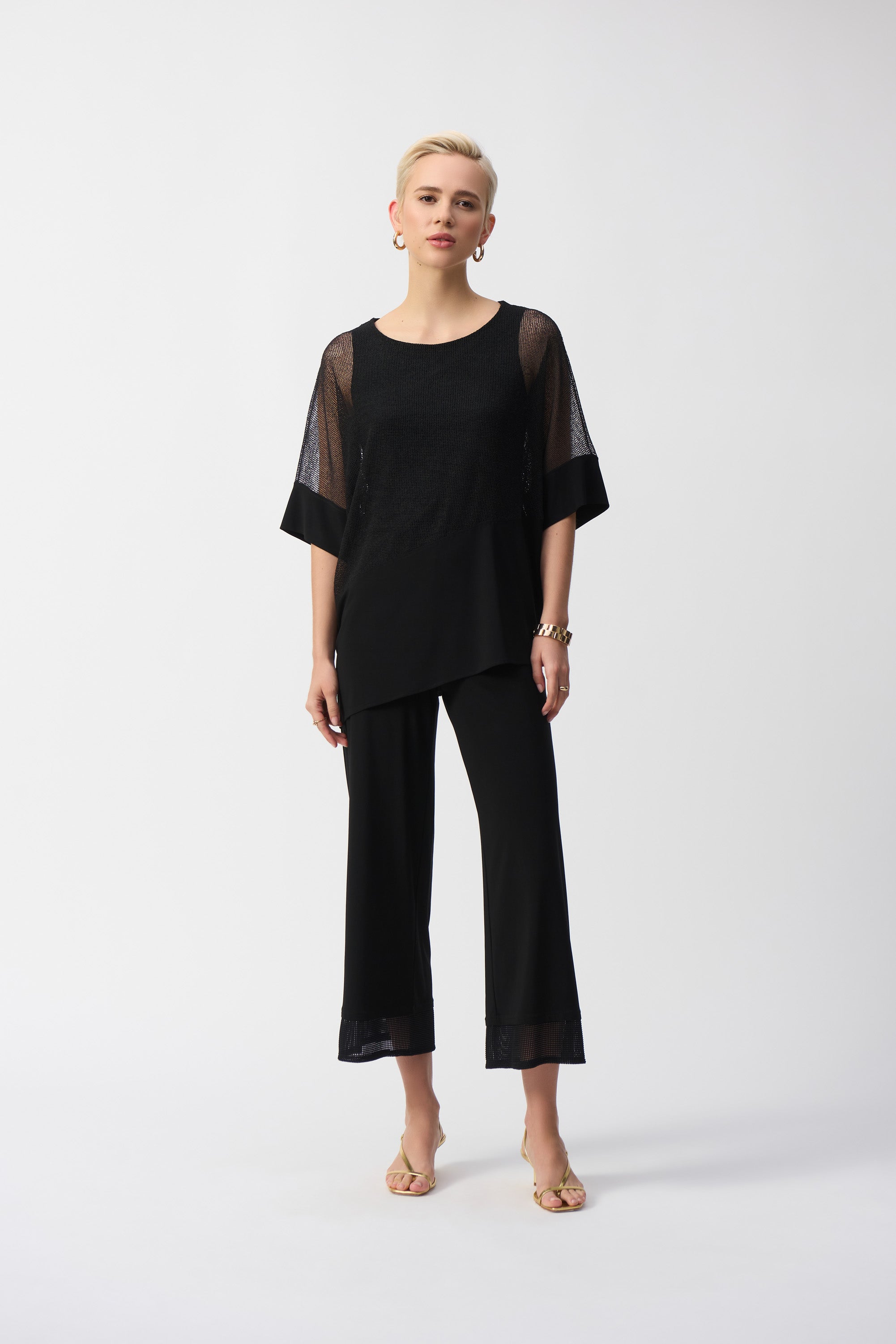 Woman wearing a black mesh cocoon top with asymmetrical hem and matching black pants, standing in full view.