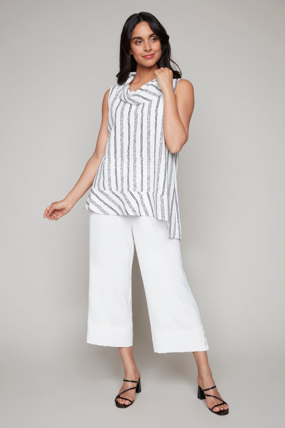 Woman wearing a white sleeveless cowl neck top with black vertical stripes and side slits, paired with wide-leg white pants and black heeled sandals.
