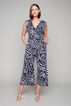 Woman wearing a sleeveless navy and white abstract print jumpsuit with a V-neckline, tie waist, and wide cropped legs, styled with white strappy heels.