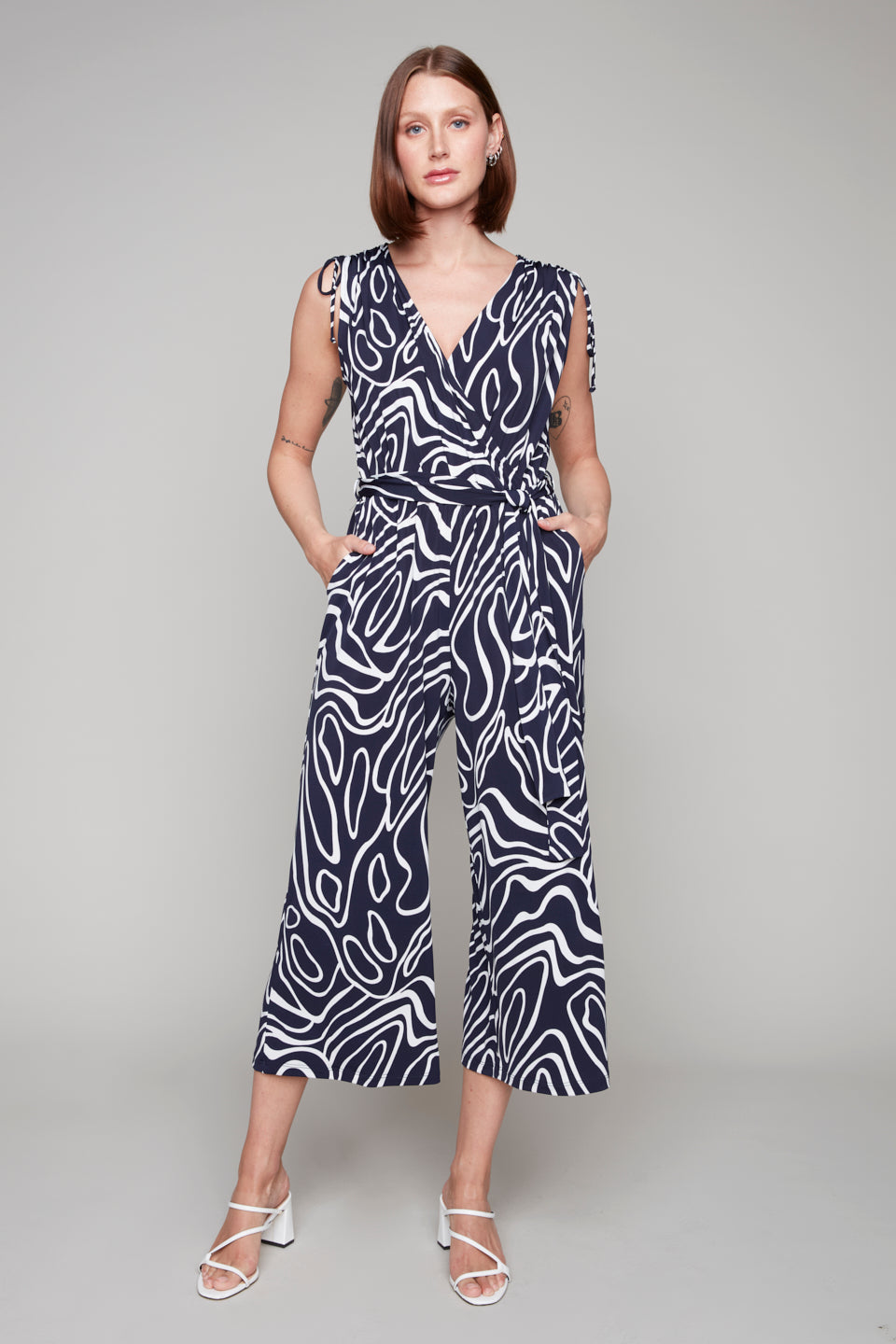 Woman wearing a sleeveless navy and white abstract print jumpsuit with a V-neckline, tie waist, and wide cropped legs, styled with white strappy heels.