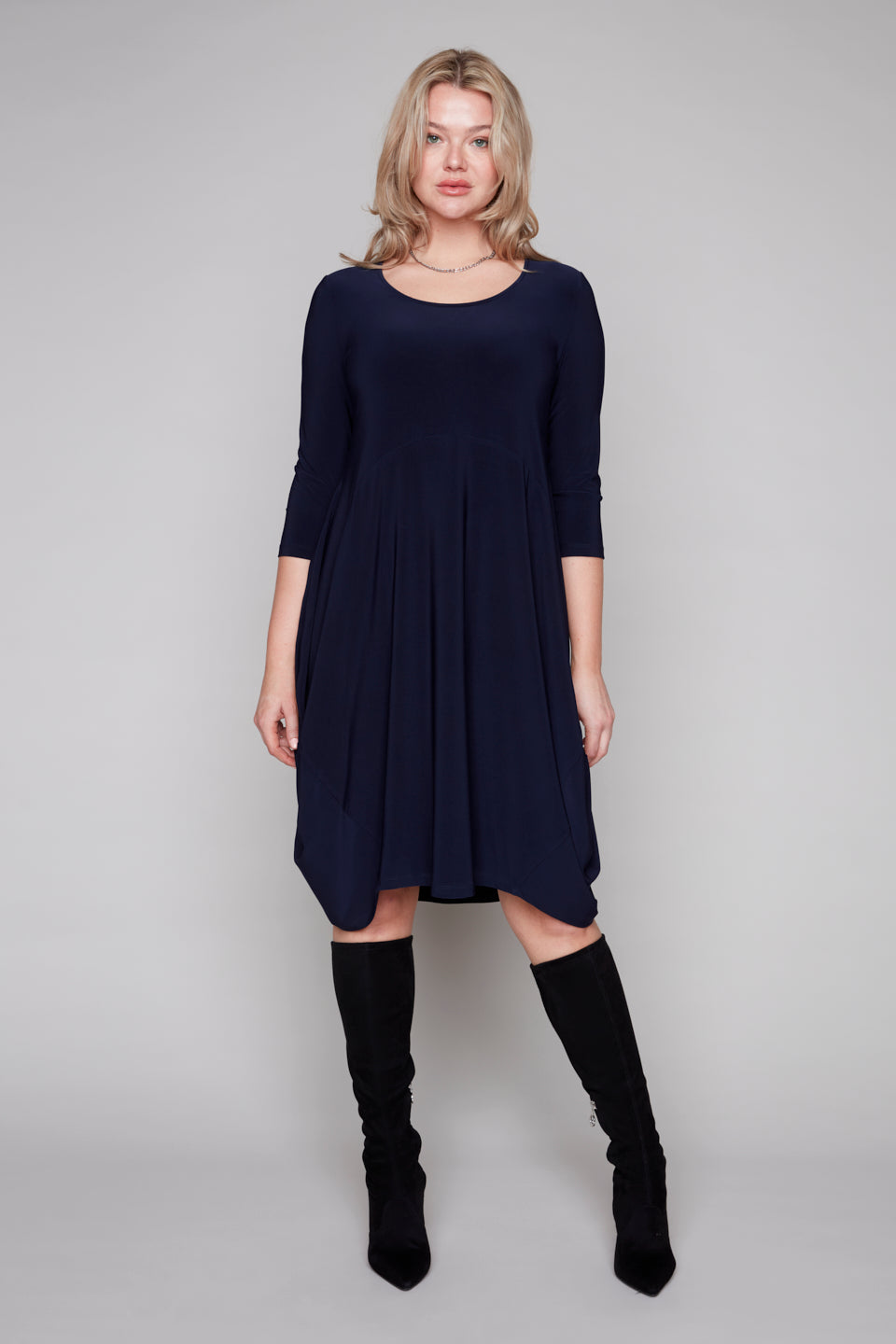 Woman wearing a navy blue dress with long sleeves and black knee-high boots on a gray background