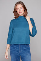 Woman wearing a blue turtleneck sweater and jeans against a gray background