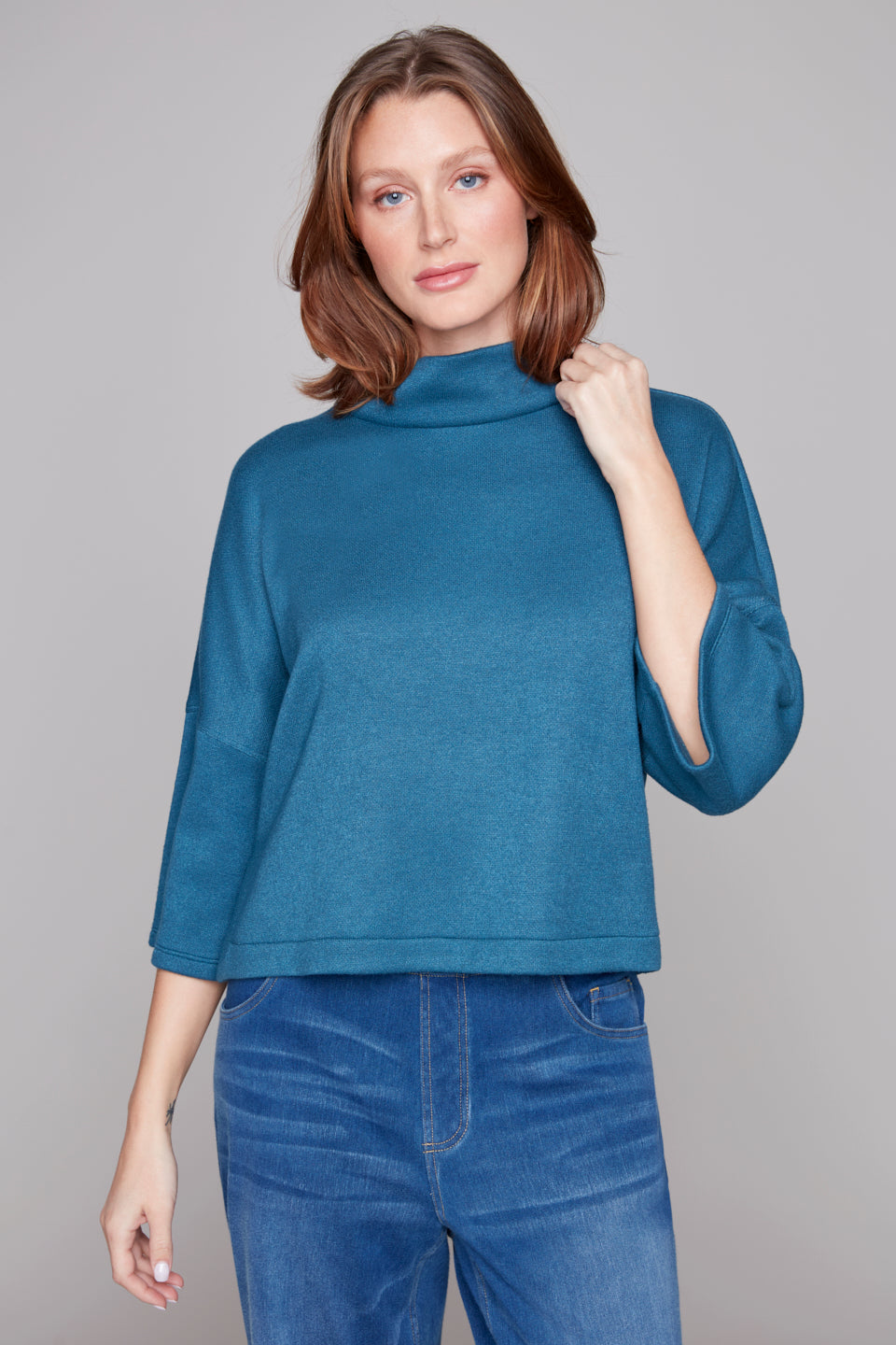 Woman wearing a blue turtleneck sweater and jeans against a gray background