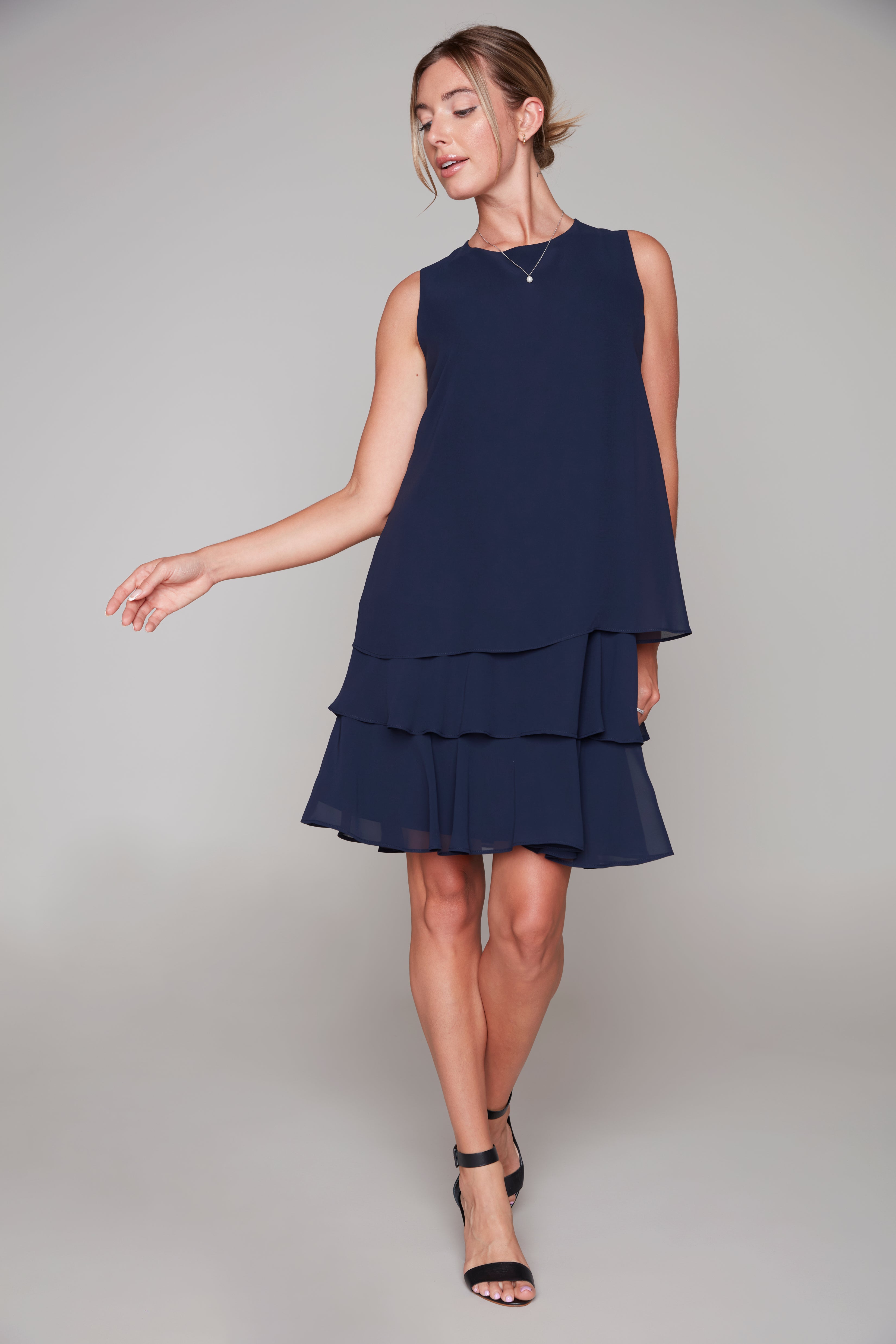 Model wearing a sleeveless navy layered chiffon dress with a round neckline and tiered ruffle hem, styled with black strappy heels.