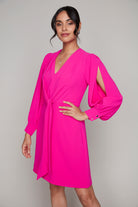 Woman wearing a vibrant fuchsia long-sleeve dress featuring a V-neckline, gathered front tie detail, and dramatic shoulder slits with buttoned cuffs.







