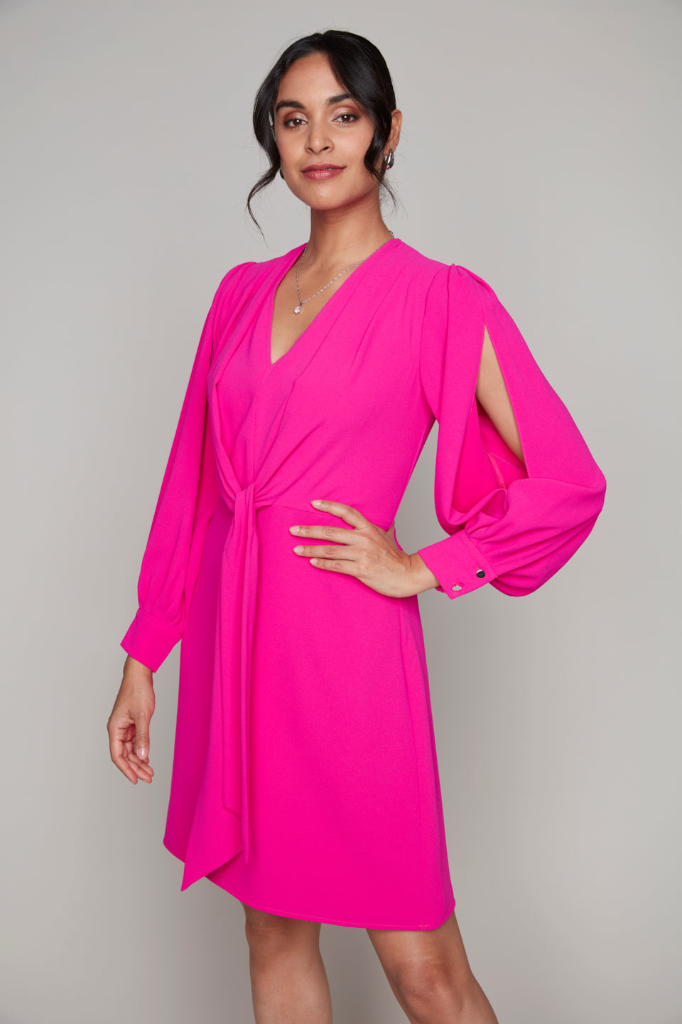 Woman wearing a vibrant fuchsia long-sleeve dress featuring a V-neckline, gathered front tie detail, and dramatic shoulder slits with buttoned cuffs.







