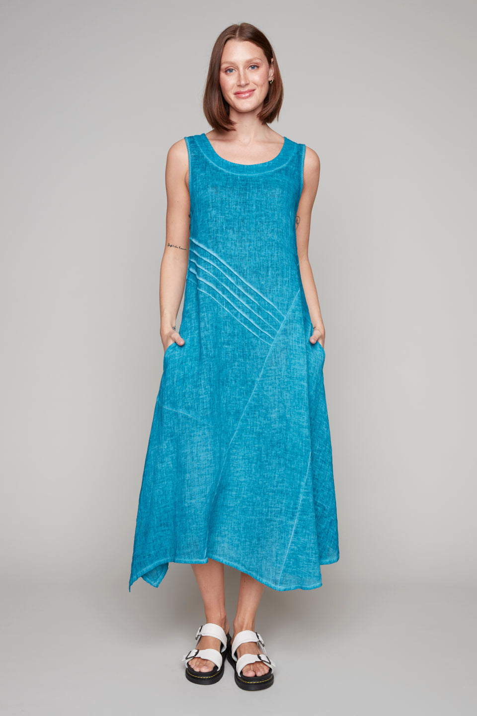 Woman wearing a sleeveless aqua blue midi dress with diagonal pleat detail and asymmetrical hemline.