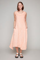 Woman wearing a sleeveless salmon pink midi dress with diagonal pleat detail and asymmetrical hemline.