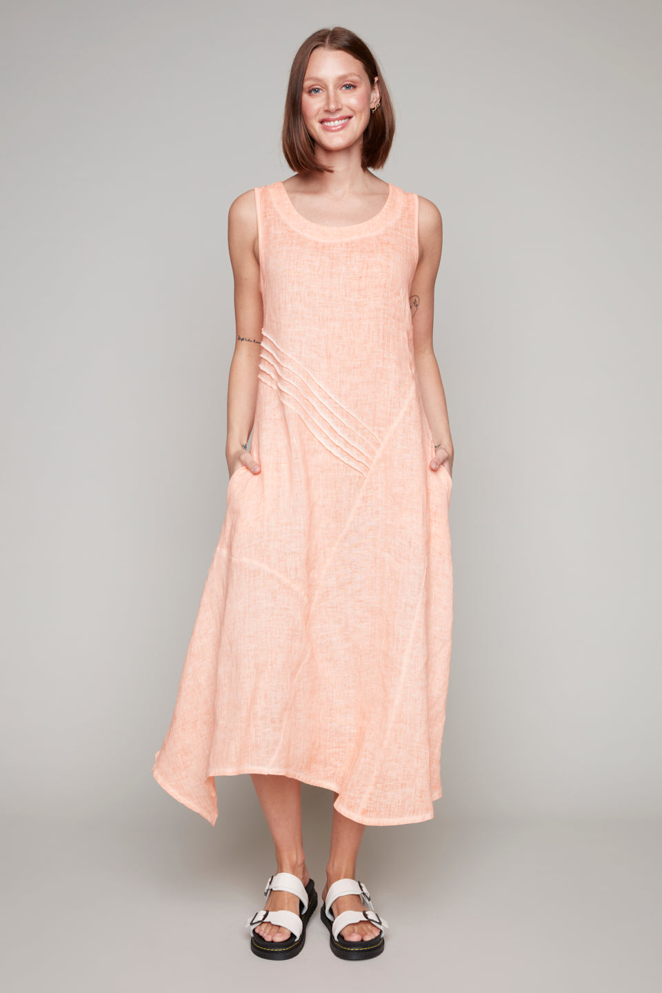 Woman wearing a sleeveless salmon pink midi dress with diagonal pleat detail and asymmetrical hemline.