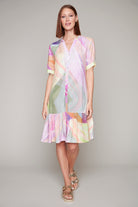 Woman wearing a colorful abstract-print button-down shirt dress with short cuffed sleeves and a tiered hem, styled with gold sandals.