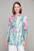 Woman wearing a long-sleeve button-up shirt in a painterly floral print with a high-low hem and classic collar, styled with white pants.