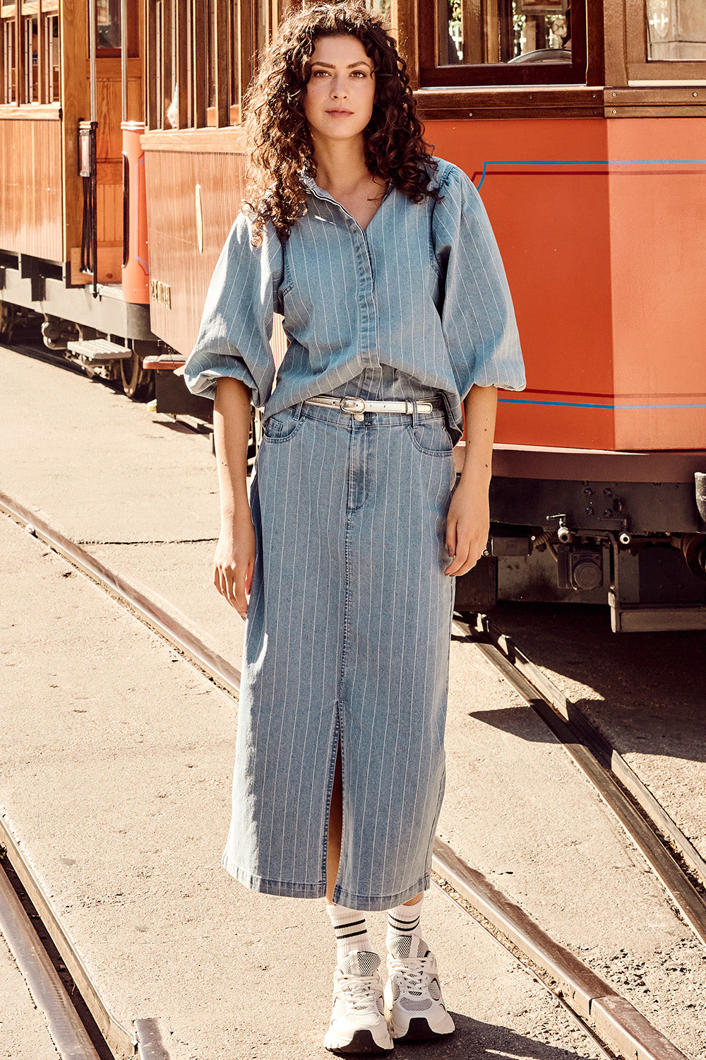 Woman standing near a vintage red tram wearing a light blue denim pinstripe blouse with puff sleeves and a matching high-waisted midi skirt with front slit, styled with a white belt and sneakers.