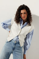 Woman wearing a light blue and white pinstripe blouse tucked into light denim jeans, layered with a sleeveless white knit vest featuring gold buttons.