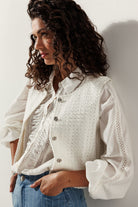 Side view of a woman wearing a textured white vest with crystal buttons over a white blouse with ruffle and lace details, paired with blue pinstripe denim.