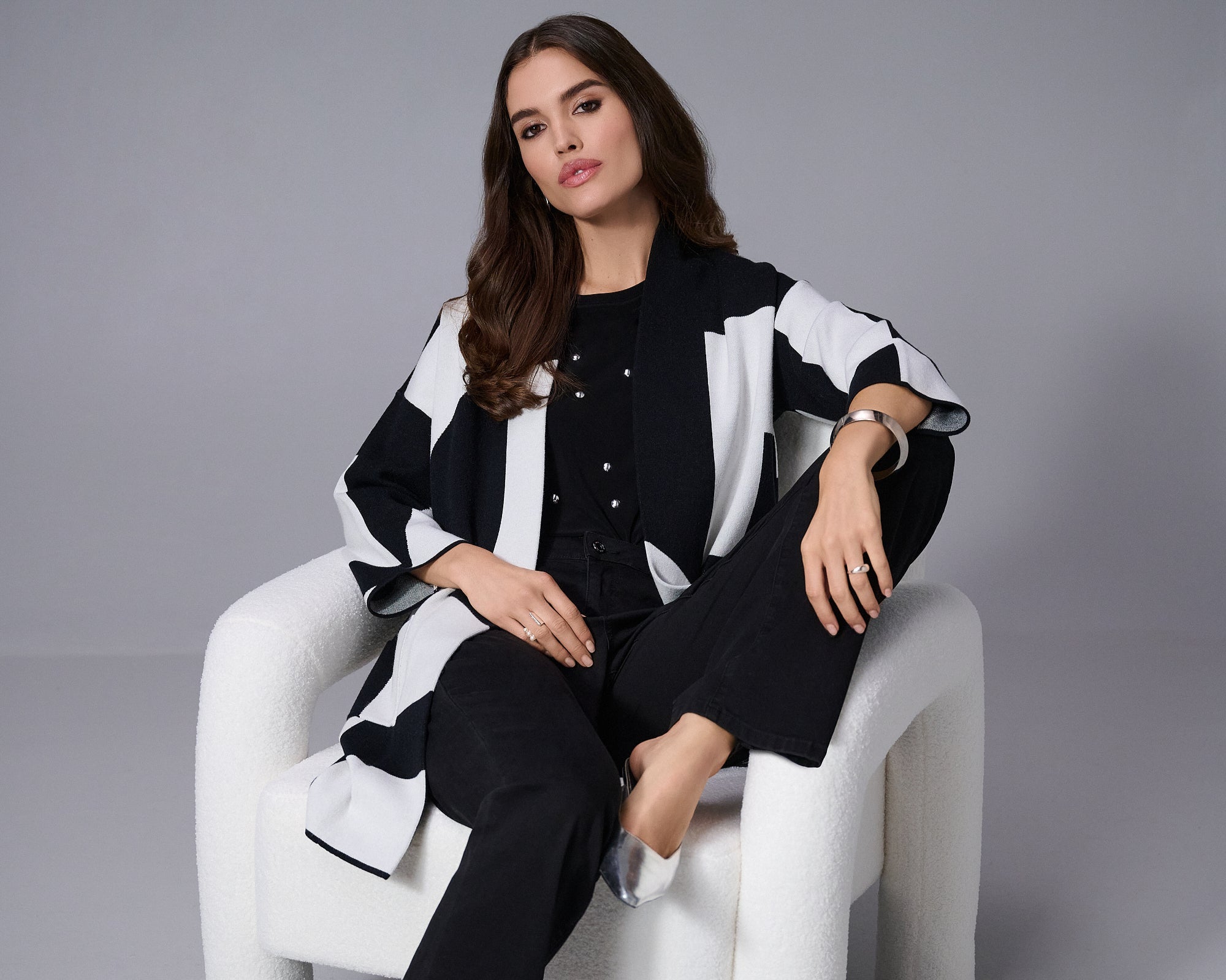 Woman sitting on a white chair wearing a black and white outfit against a gray background
