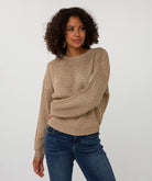 Woman wearing a beige sweater and blue jeans against a white background arm across stomach