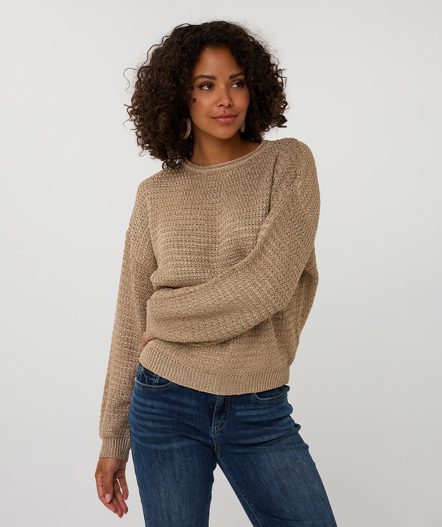 Woman wearing a beige sweater and blue jeans against a white background arm across stomach