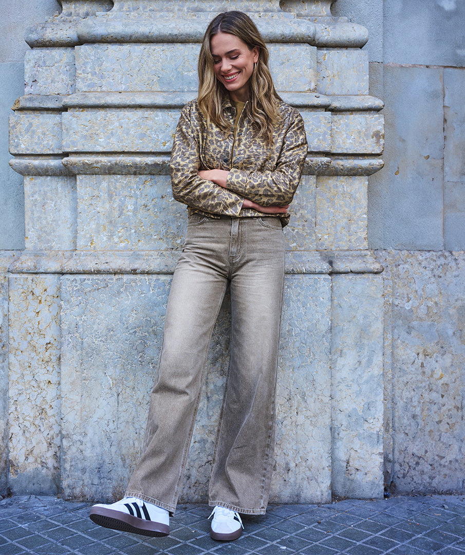 Woman wearing a patterned shirt and beige pants leaning against a stone wall.