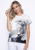 Woman wearing a short sleeve printed top with oversized black floral line art and contrast black-and-white stripe detailing on back paired with white pants.