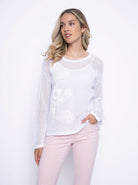 Woman wearing a long sleeve white mesh top with floral appliqué on the front, styled with pale pink jeans.