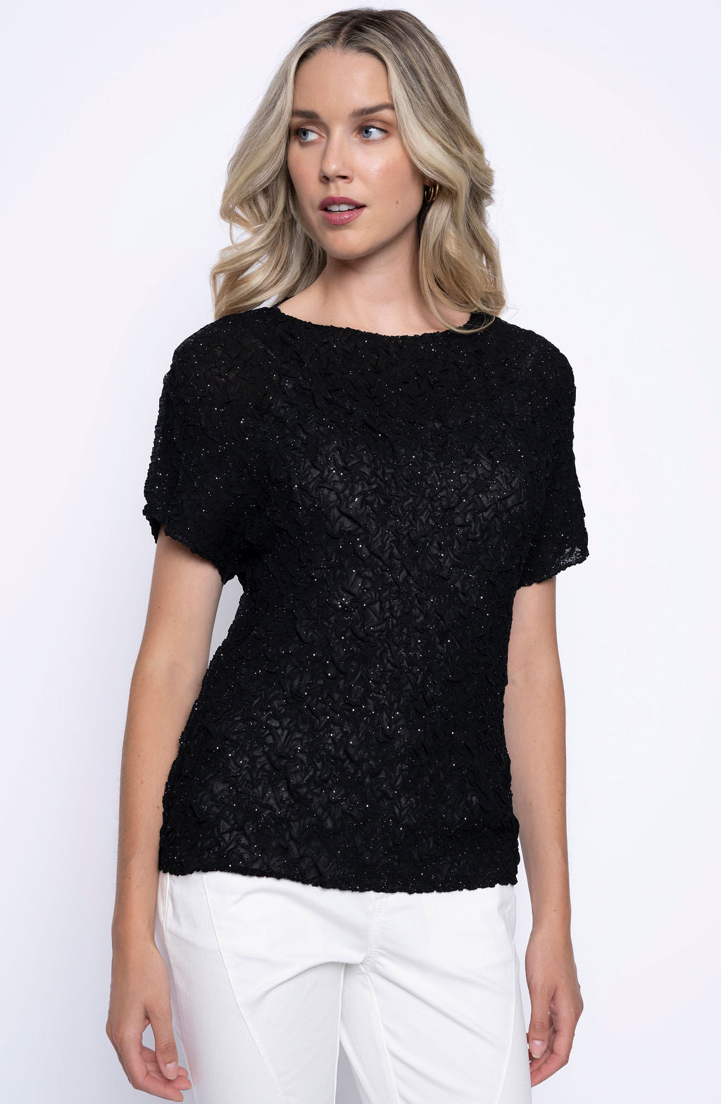 Woman wearing short sleeve black textured pleated top with subtle shimmer, paired with white pants.