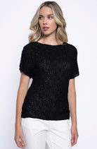 Woman wearing short sleeve black textured pleated top with subtle shimmer, paired with white pants.