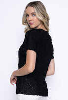 Woman showing the side view of the black pleated top with soft texture and shine, styled with white bottoms.