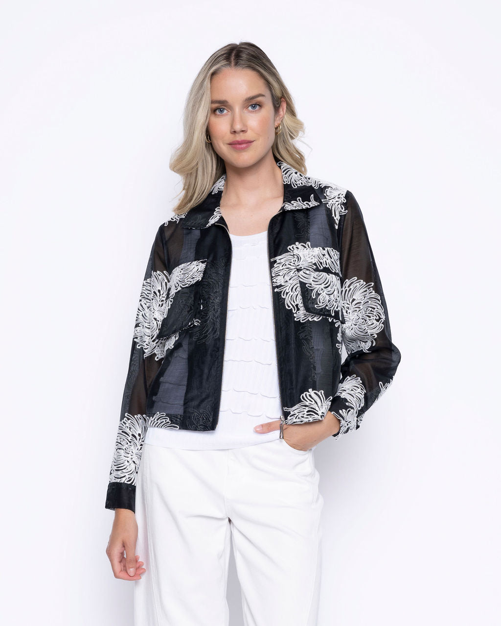 Woman wearing a sheer black zip-front cropped jacket with white floral embroidery over a white top and white pants.