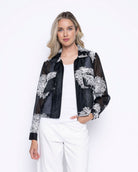 Woman wearing a sheer black zip-front cropped jacket with white floral embroidery over a white top and white pants.