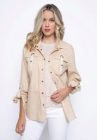 Woman wearing a beige button-front jacket with front flap pockets and grommet detailing, styled open over a light top with white pants.