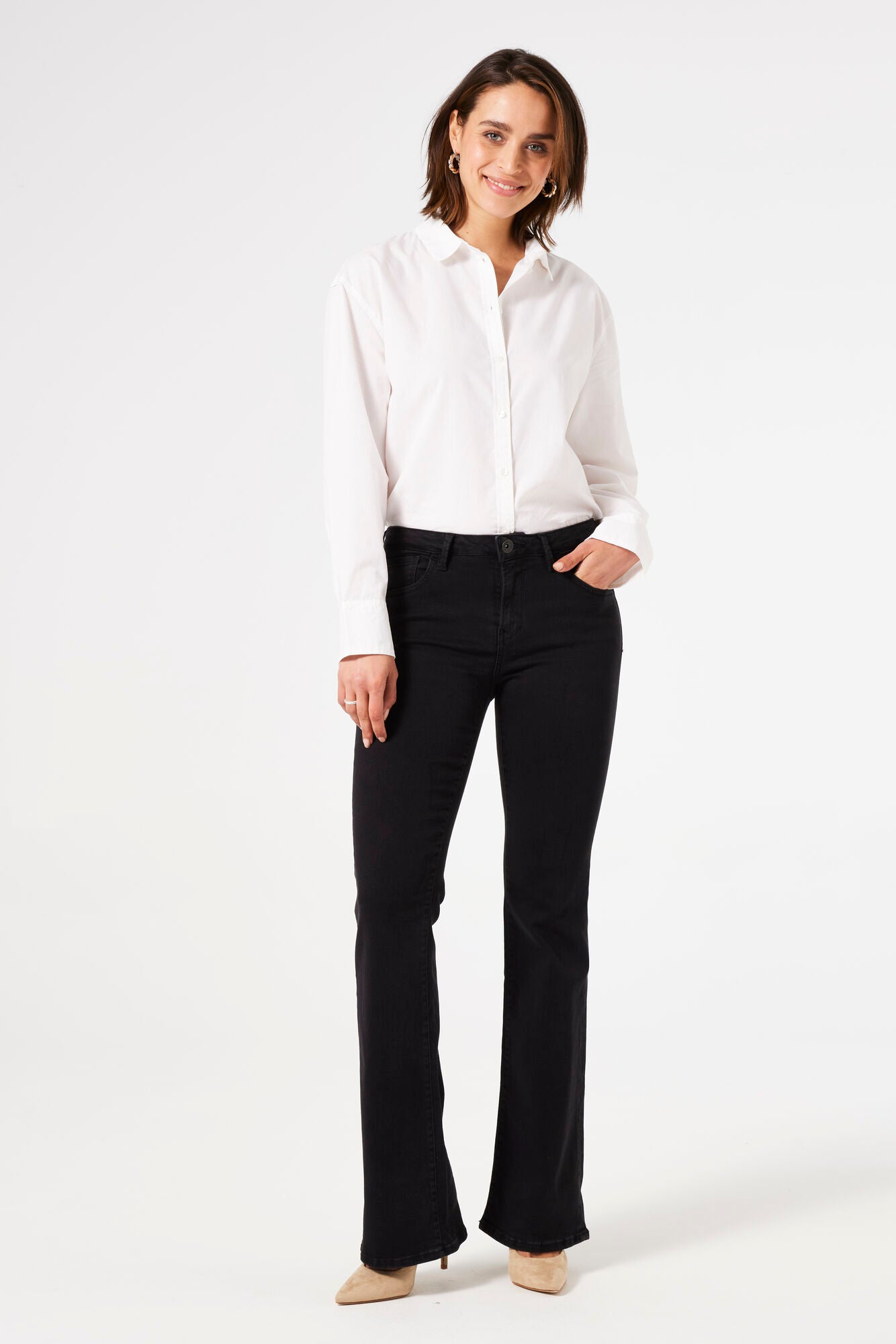 Front-angled view of model wearing black bootcut jeans with one hand in her pocket, styled with hoop earrings and beige heels.