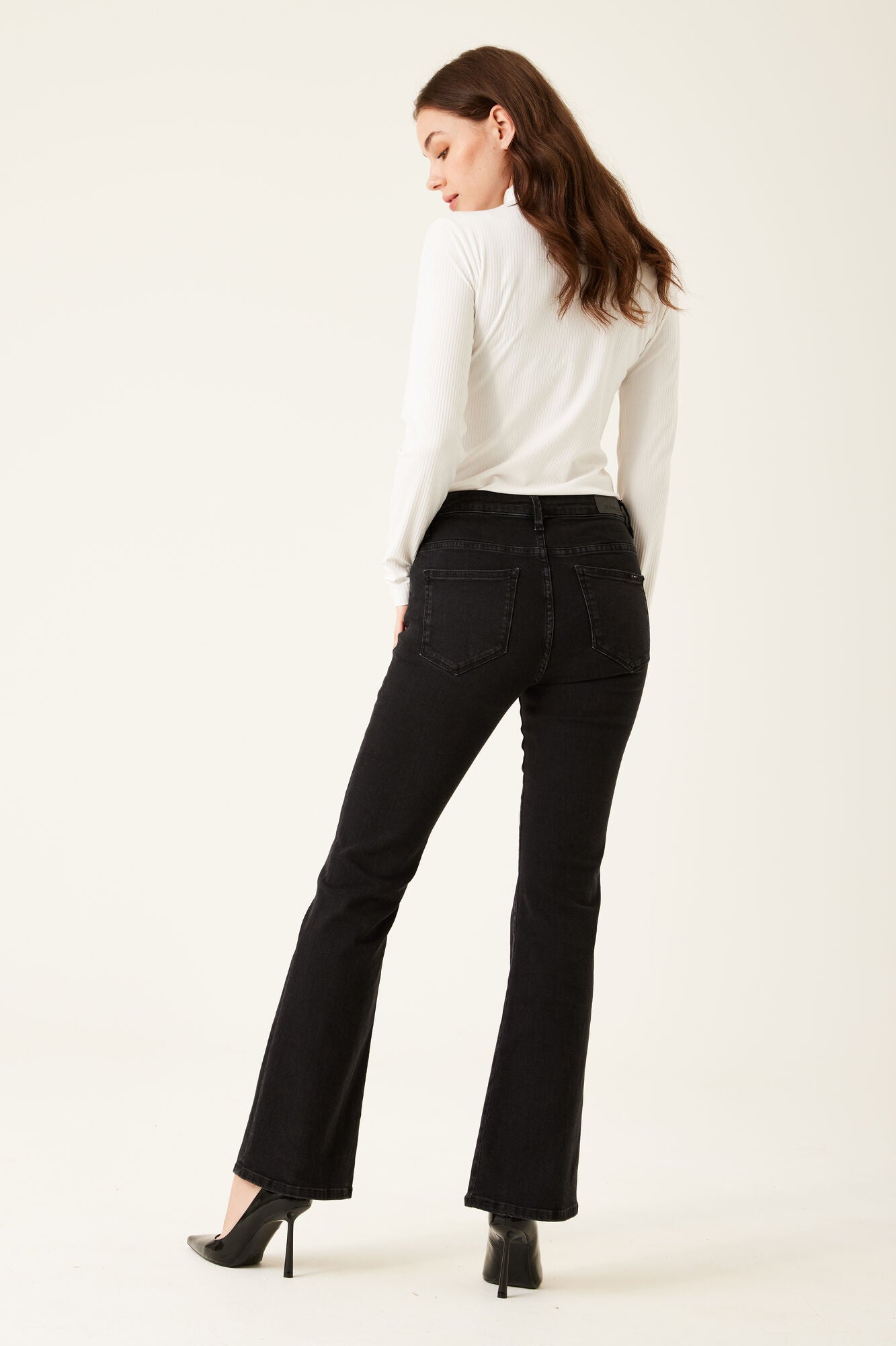 Back view of woman wearing black bootcut jeans with a fitted white long-sleeve top and black heels.
