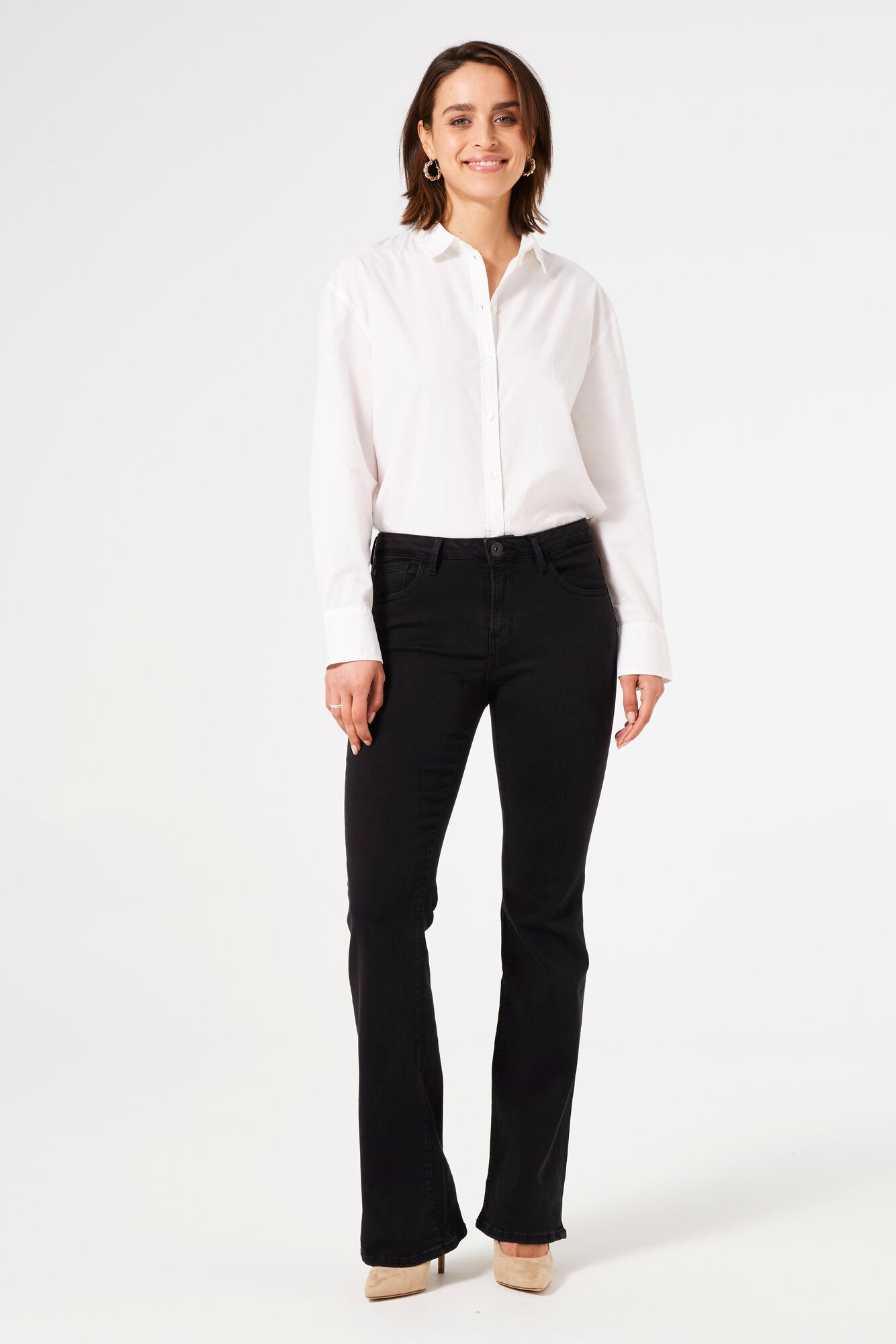 Model wearing black bootcut jeans with a crisp white shirt and beige heels, smiling directly at camera.