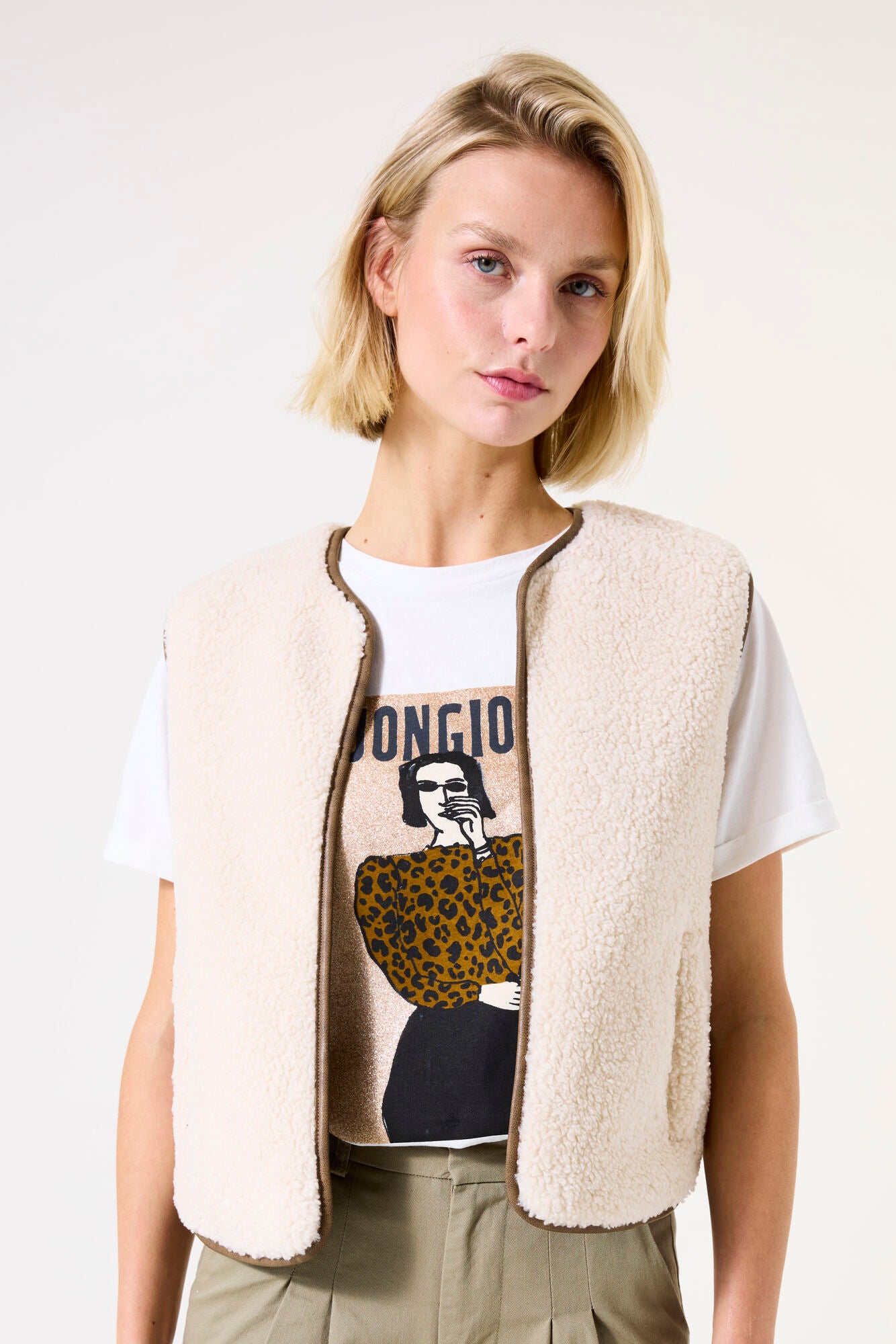 Woman wearing a white t-shirt with graphic design and a beige vest on a plain background