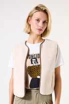 Woman wearing a white t-shirt with graphic design and a beige vest on a plain background