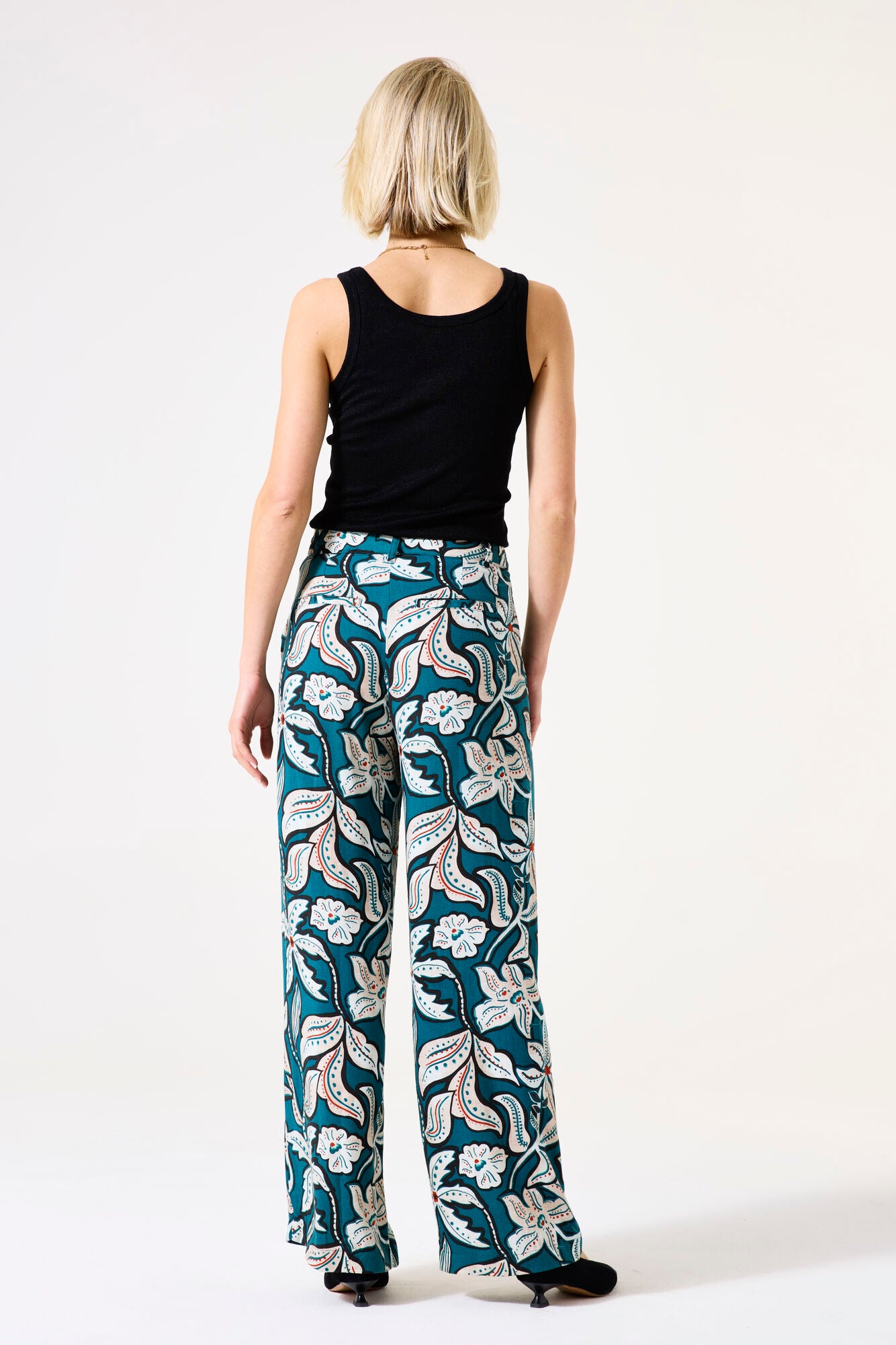 Back View of Woman wearing a black sleeveless top and floral pants on a white background