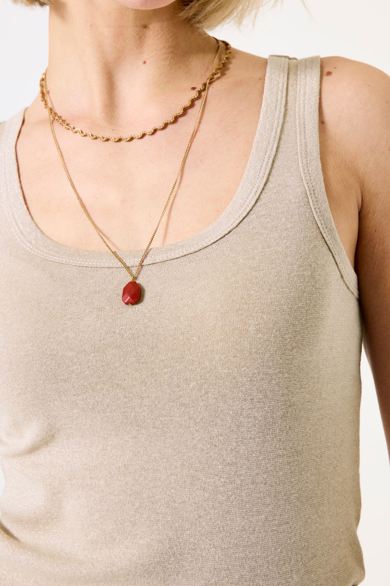 Person wearing a beige tank top with a gold necklace featuring a red pendant.