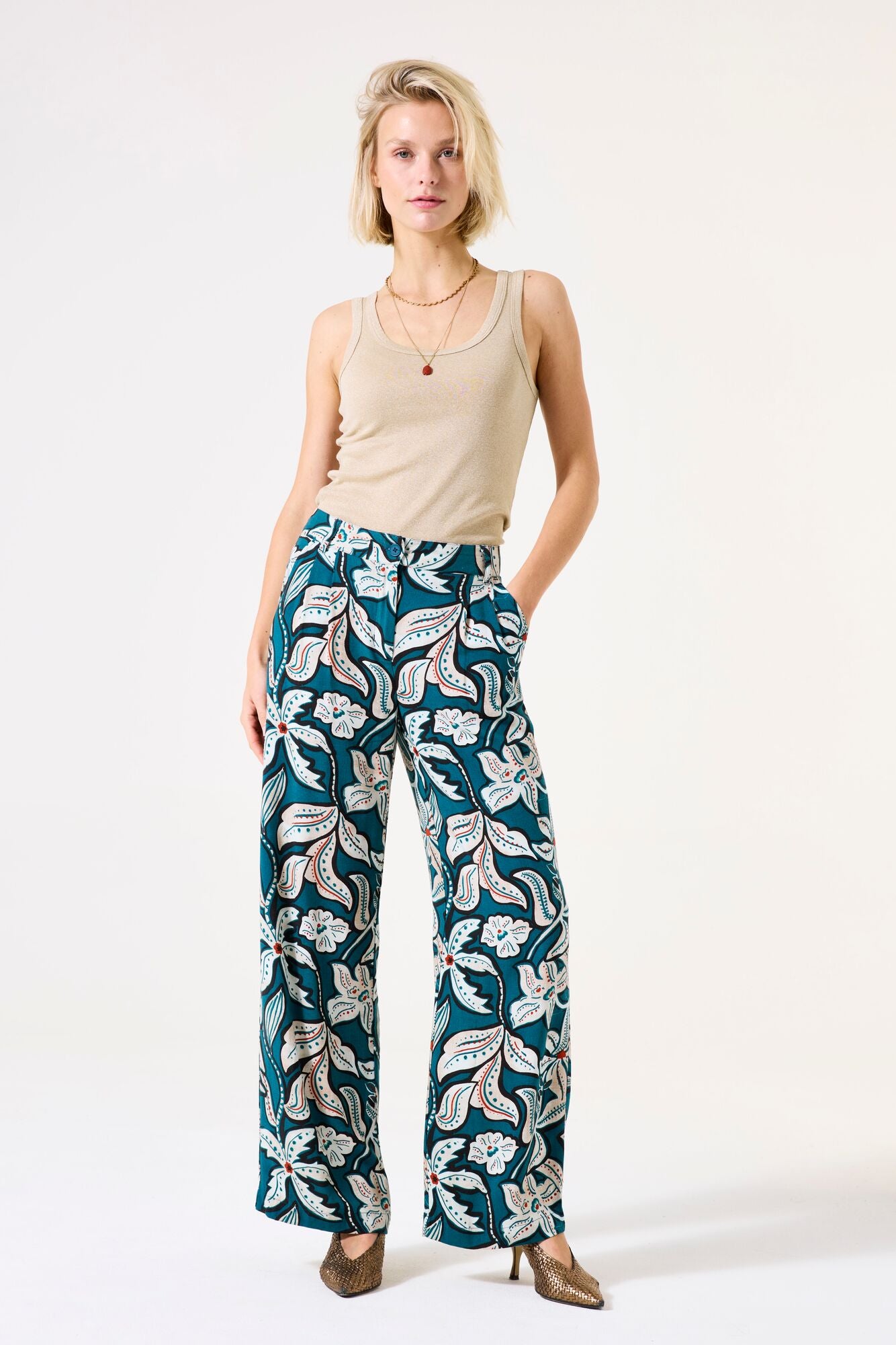 Woman wearing a beige tank top and teal floral pants on a white background